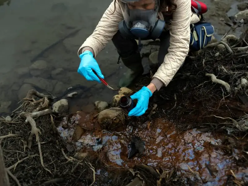 Scientist collecting soil samples wearing protective gear near water.