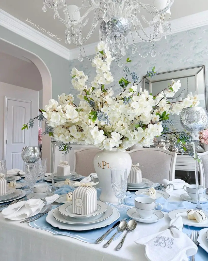 Elegant dining table with crystal chandelier, large white floral centerpiece, and blue accents.