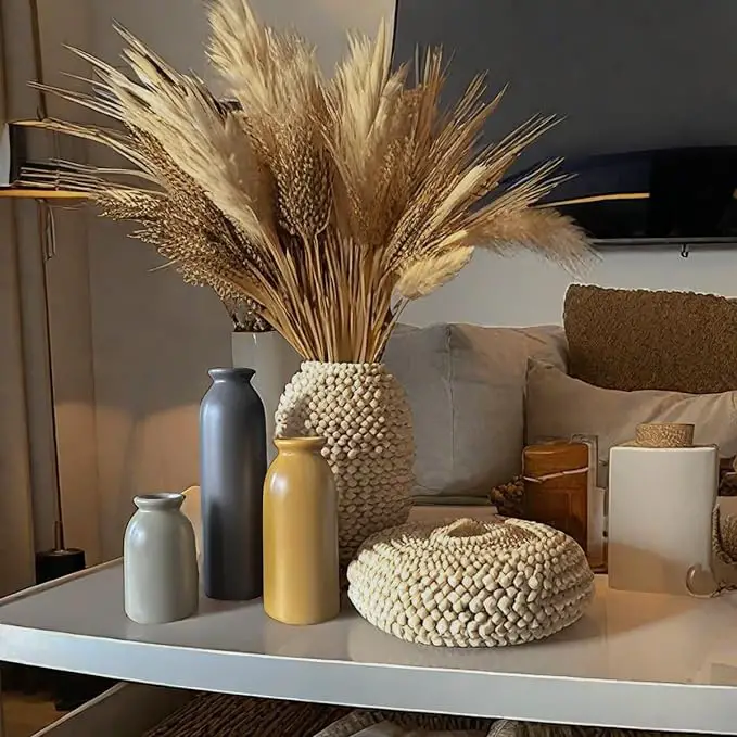 A modern living room features a white coffee table adorned with a woven vase holding dried pampas grass, accompanied by various ceramic vases in shades of gray, yellow, and white.