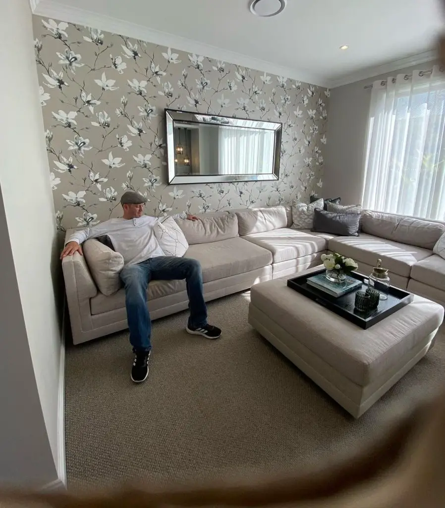 Man relaxing on beige sectional sofa in living room with floral wallpaper.