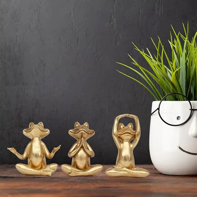 Three small golden frog figurines in yoga poses are placed on a wooden surface next to a white face-shaped planter with glasses, containing green grass-like plants