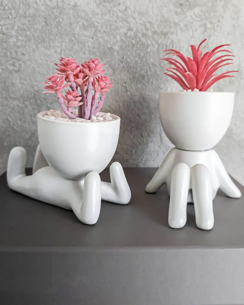 Two small, white ceramic planters shaped like minimalist human figures in different poses hold pink cactus and air plant.