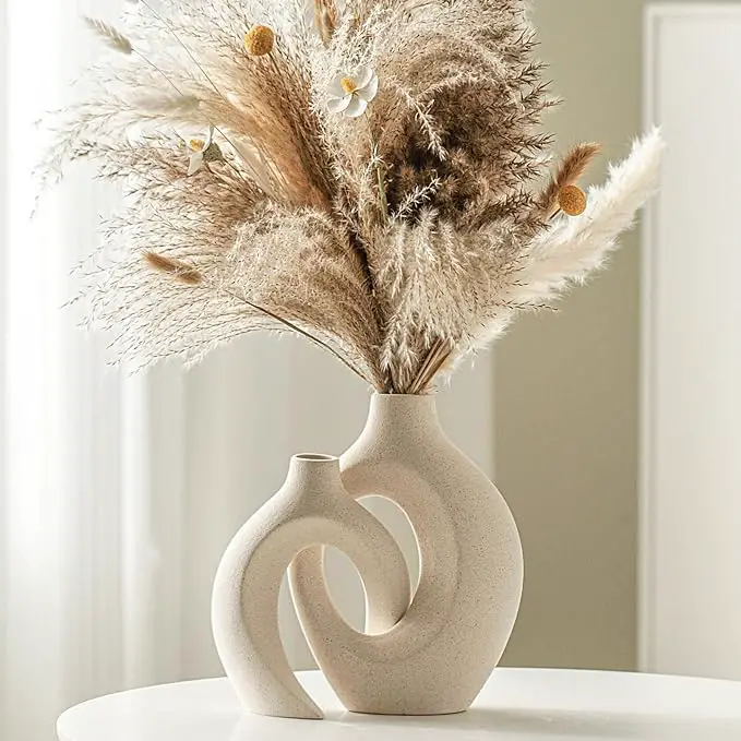 A modern beige ceramic vase with a unique intertwined design, holding an arrangement of dried pampas grass and small white flowers, placed on a clean white surface with a light background.