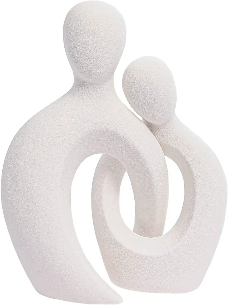 A minimalist white sculpture depicting two abstract, faceless figures: one taller with an arched body and the other smaller, leaning against the taller figure.