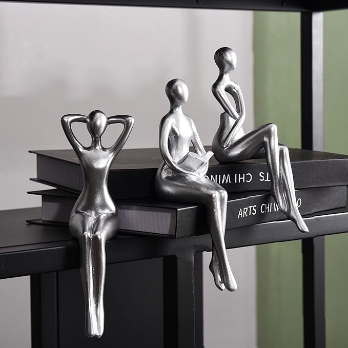 Three modern silver figurines sit on the edge of a black shelf, each with a distinct pose.