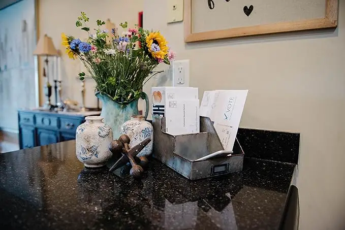 A kitchen counter holds a metal organizer with mail, a ceramic vase with a bouquet of flowers including sunflowers, two decorative ceramic jars, and a wooden trivet.