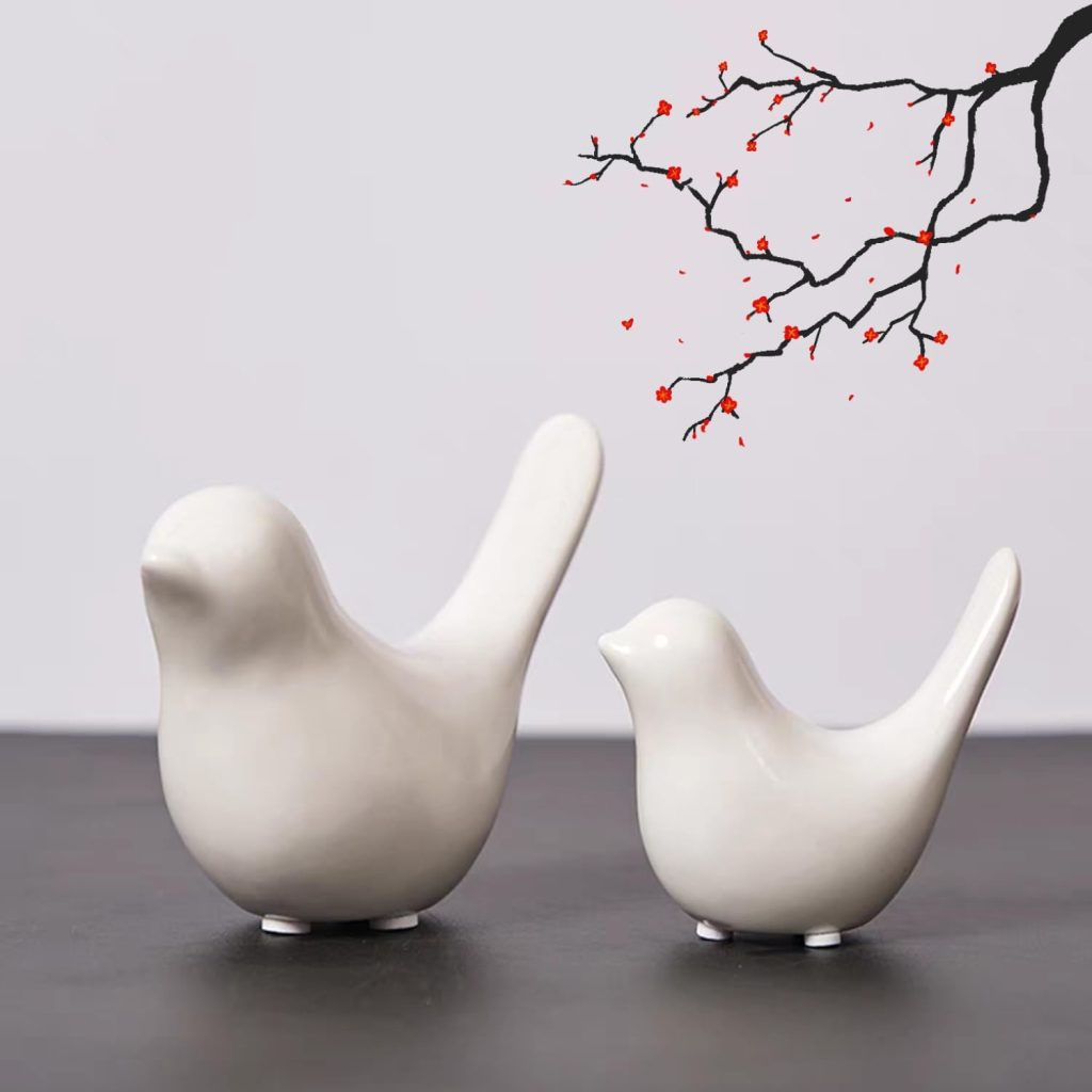 Two white ceramic bird sculptures are placed on a dark surface with a plain light gray background.