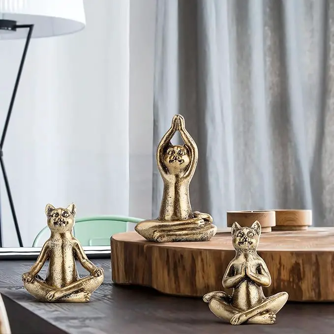 Three small gold-colored cat statues in yoga poses sit on a wooden surface in front of a cutting board.