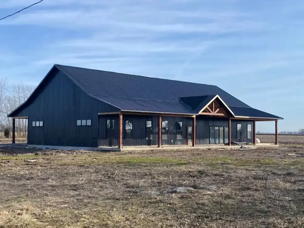 Modern black metal barndominium with timber accents and wraparound porch against rural landscape