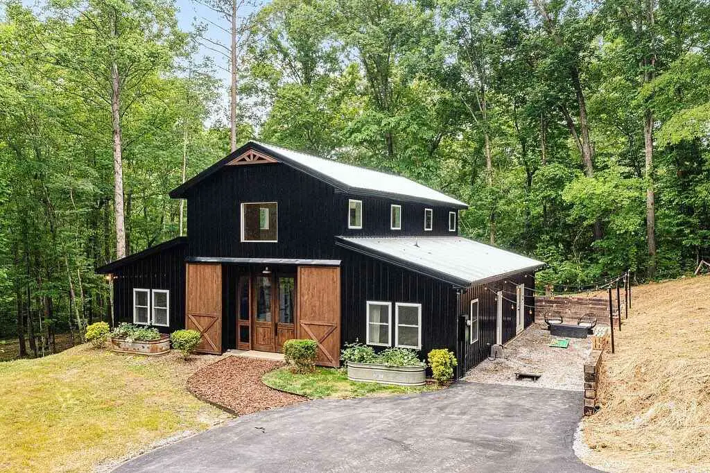 Black barndominium with wooden accents nestled in lush forest clearing with curved driveway