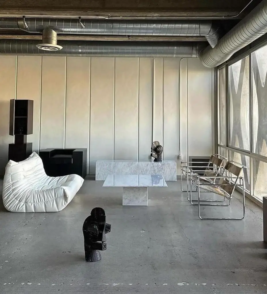 An industrial minimalist living room with exposed ductwork, concrete floor, white Togo sofa, marble coffee table, Wassily chairs, and artistic sculptures.