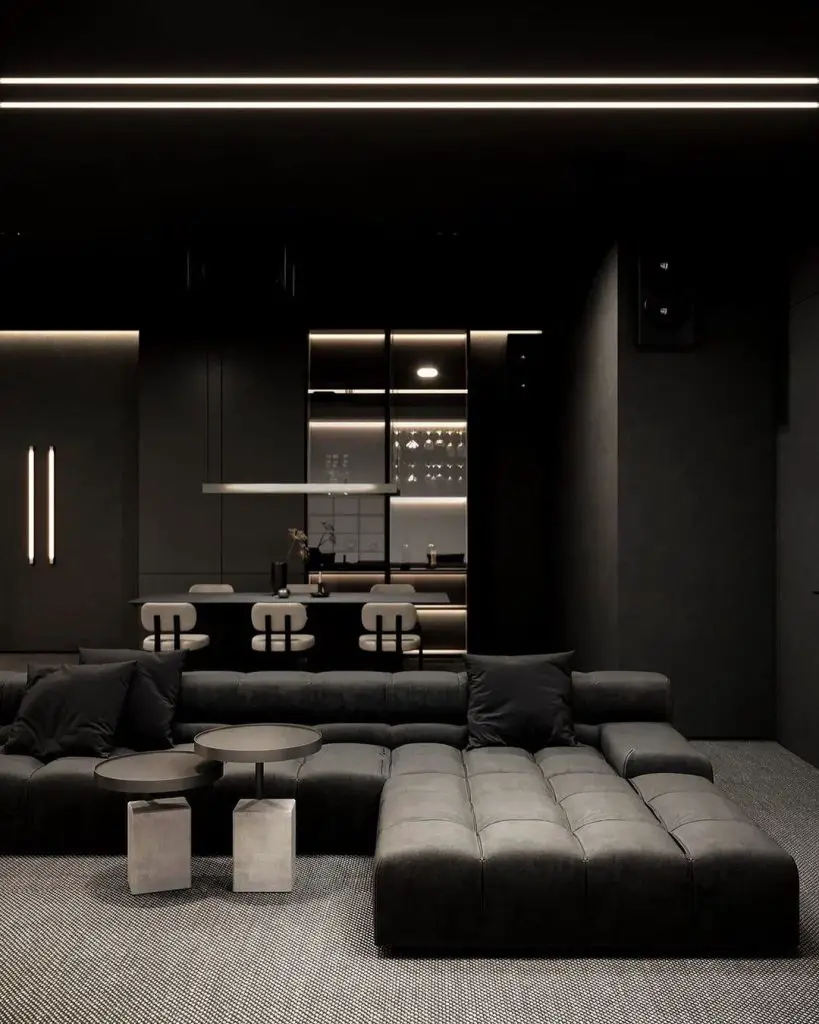 Dark living room with black sofa and illuminated kitchen beyond.