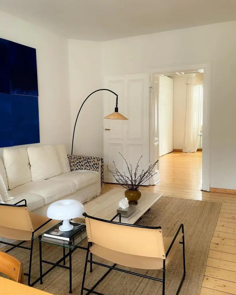 A minimalist living room with white walls, blue abstract art, white sofa, modern armchairs, and natural wood flooring.