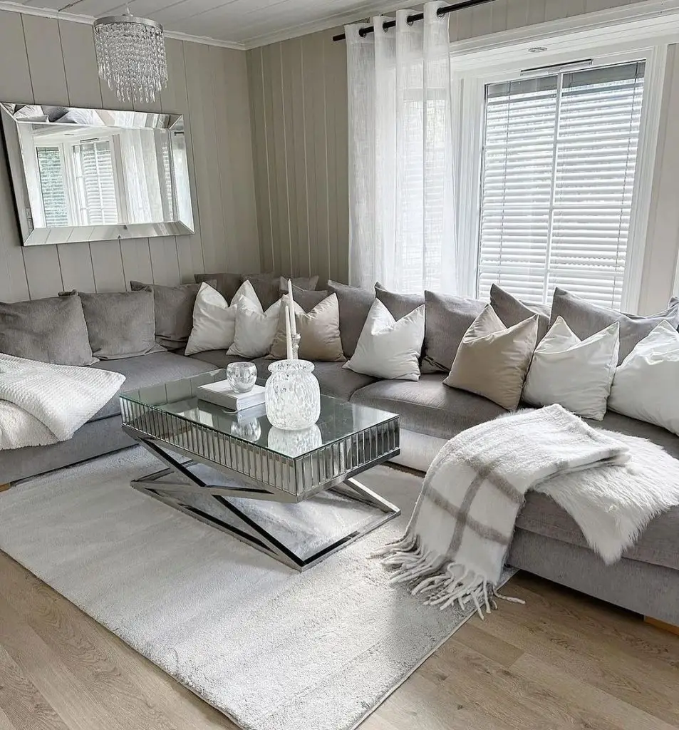 Minimalist living room with large grey sectional sofa, mirrored coffee table, crystal chandelier, and white accents.