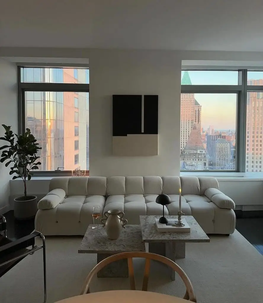 A minimalist living room with a large white sofa, marble coffee table, and floor-to-ceiling windows showcasing a city skyline at sunset.