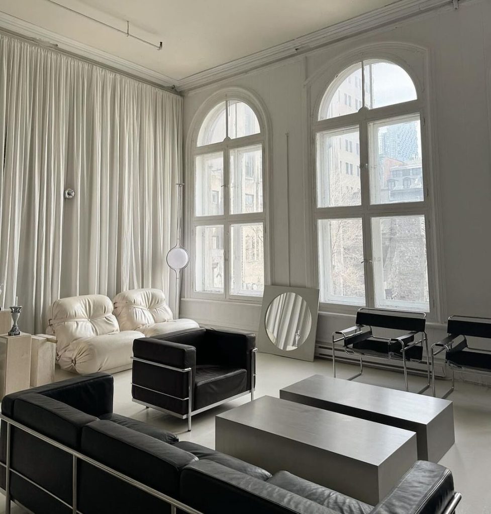 A minimalist living room with high ceilings, arched windows, white curtains, black leather chairs, a white modular sofa, and metallic cube tables.