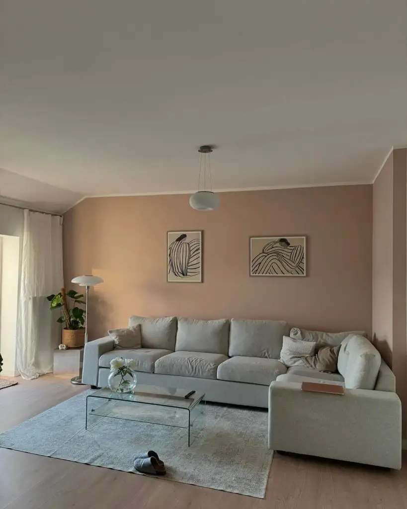 A minimalist living room with blush walls, featuring a gray L-shaped sofa, glass coffee table, abstract wall art, and modern lighting fixtures.