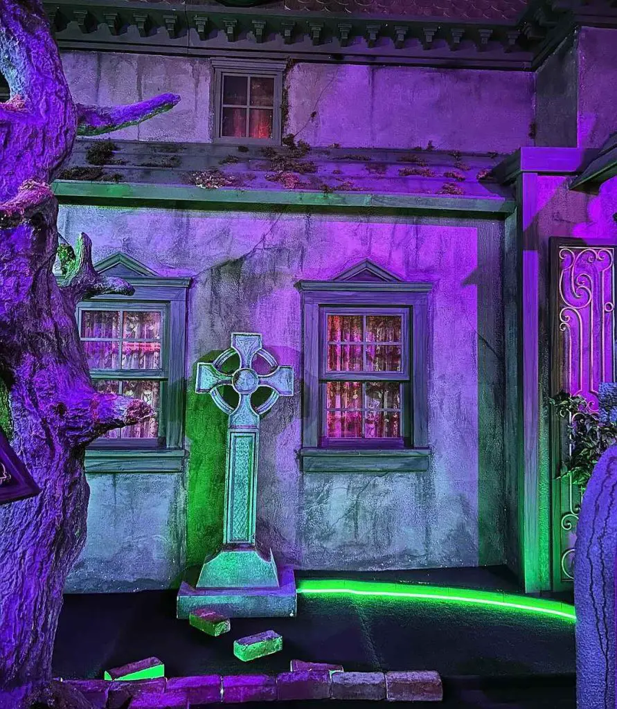 Purple-lit haunted house entrance with glowing green cross and red-lit windows.
