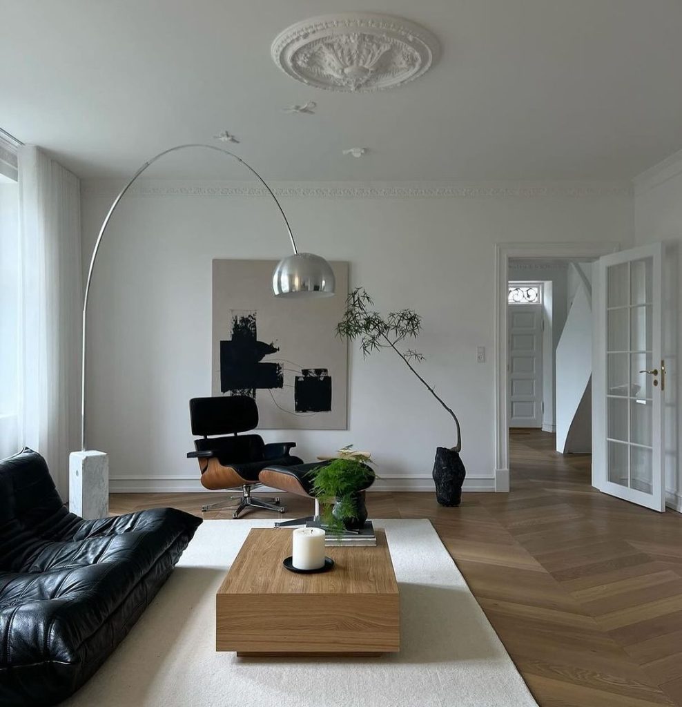 A minimalist living room with white walls, ornate ceiling medallion, black leather sofa, Eames chair, arching floor lamp, and abstract artwork.