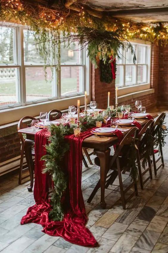 Christmas table with red velvet runner, candles, and hanging greenery in rustic room.