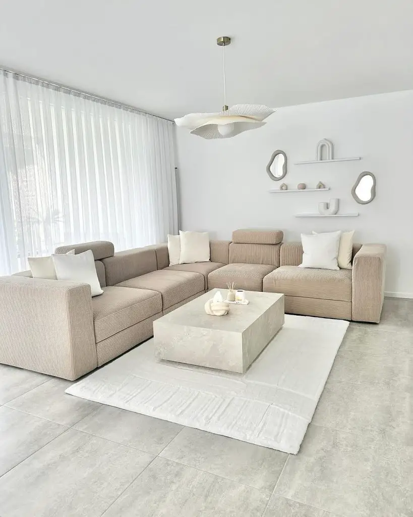 A minimalist living room with a beige sectional sofa, concrete coffee table, white pendant light, and neutral decor on floating shelves and mirrors.