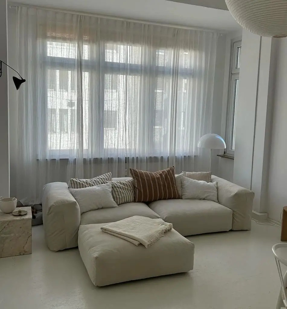 Minimalist white living room with a large sectional sofa, sheer curtains, and subtle decor elements.