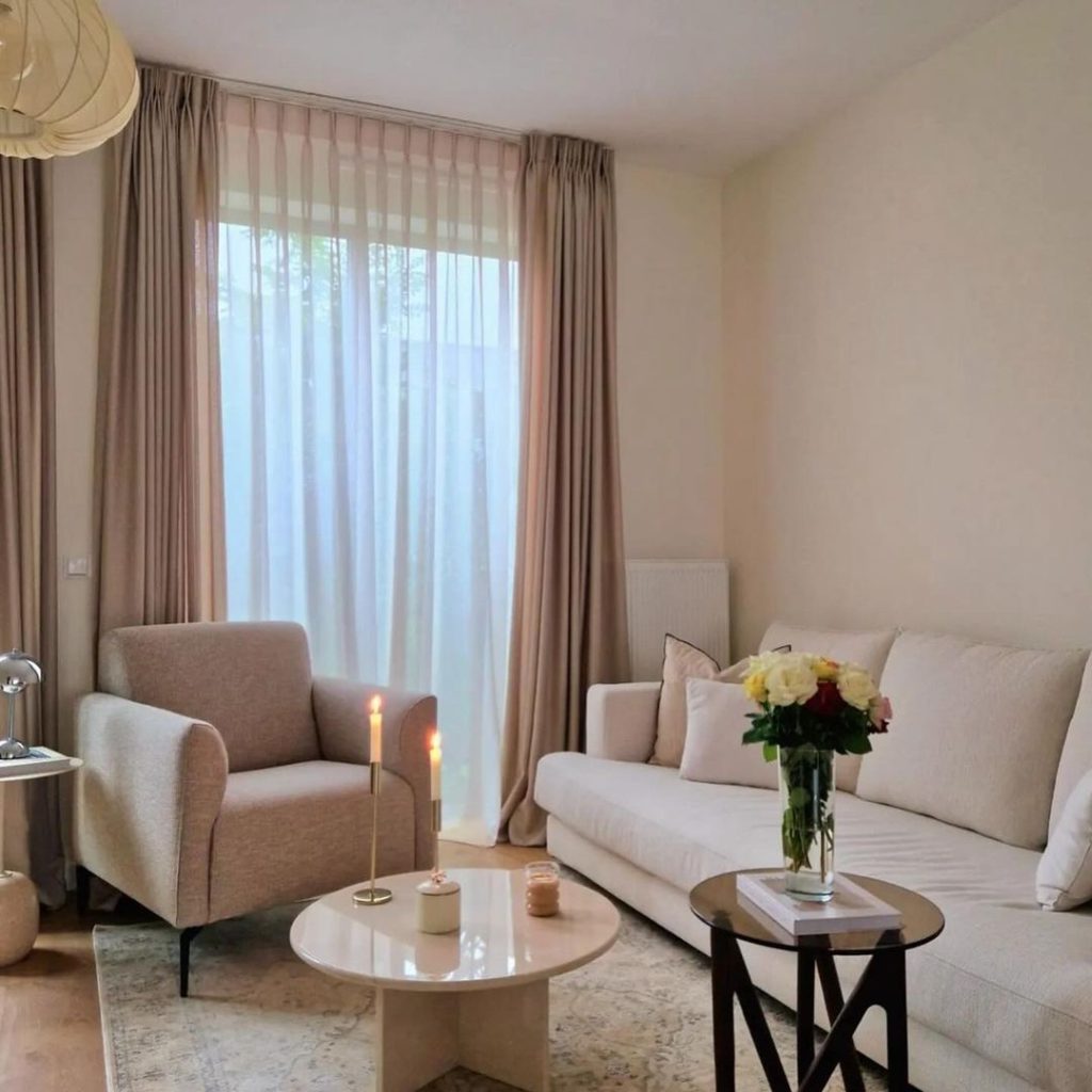 Minimalist living room with white sofa, beige armchair, round coffee table, and floor-to-ceiling curtains in neutral tones.