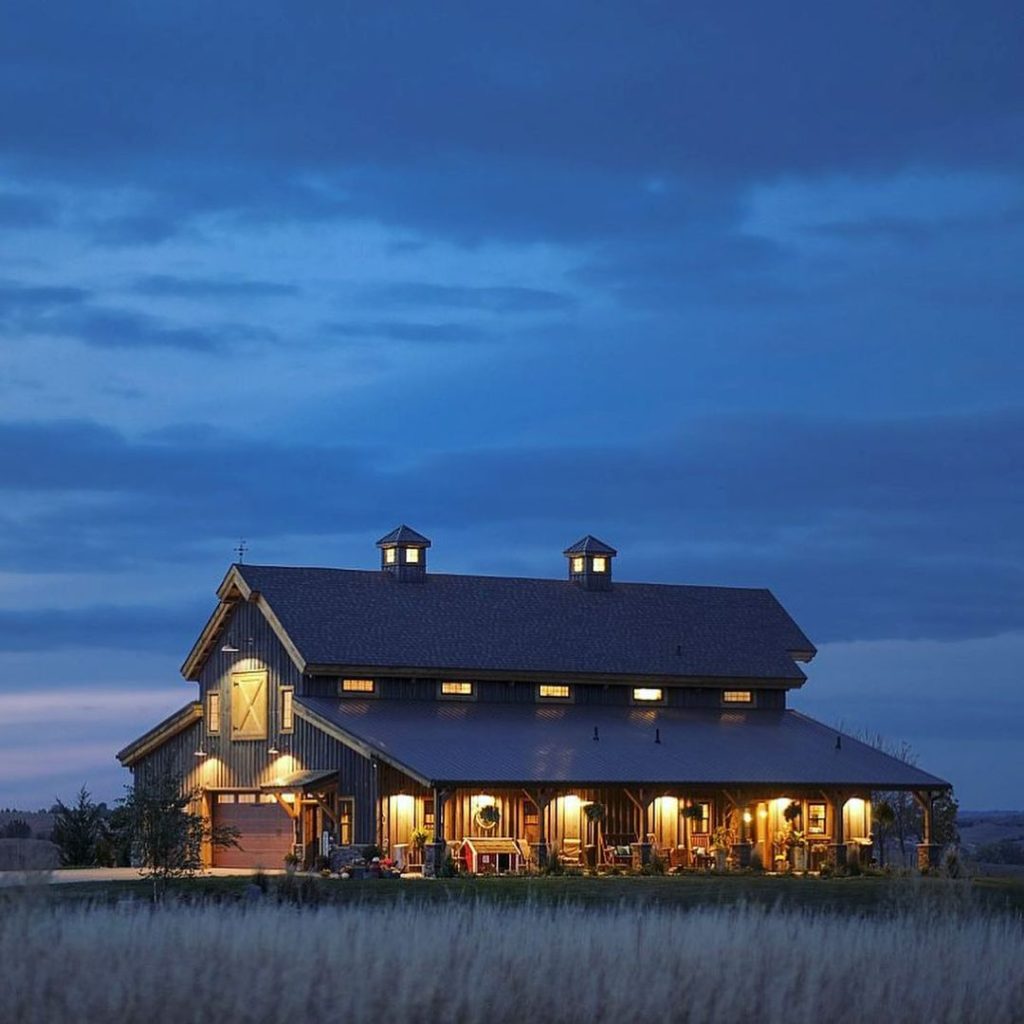 Illuminated black barn house with wraparound porch glowing against twilight prairie sky