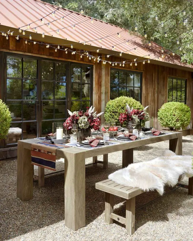 Outdoor dining setup with wooden table festoon lights and burgundy flower arrangements against rustic porch backdrop