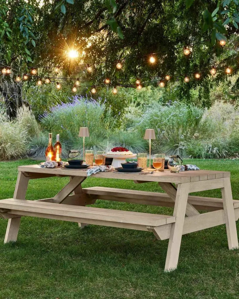 Outdoor picnic table setup with string lights overhead and garden backdrop at sunset