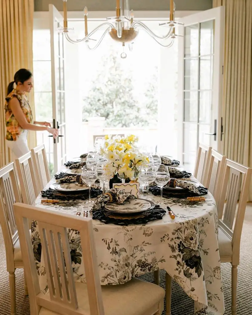Formal dining room setup with black rose tablecloth yellow daffodil centerpiece and white chairs under crystal chandelier