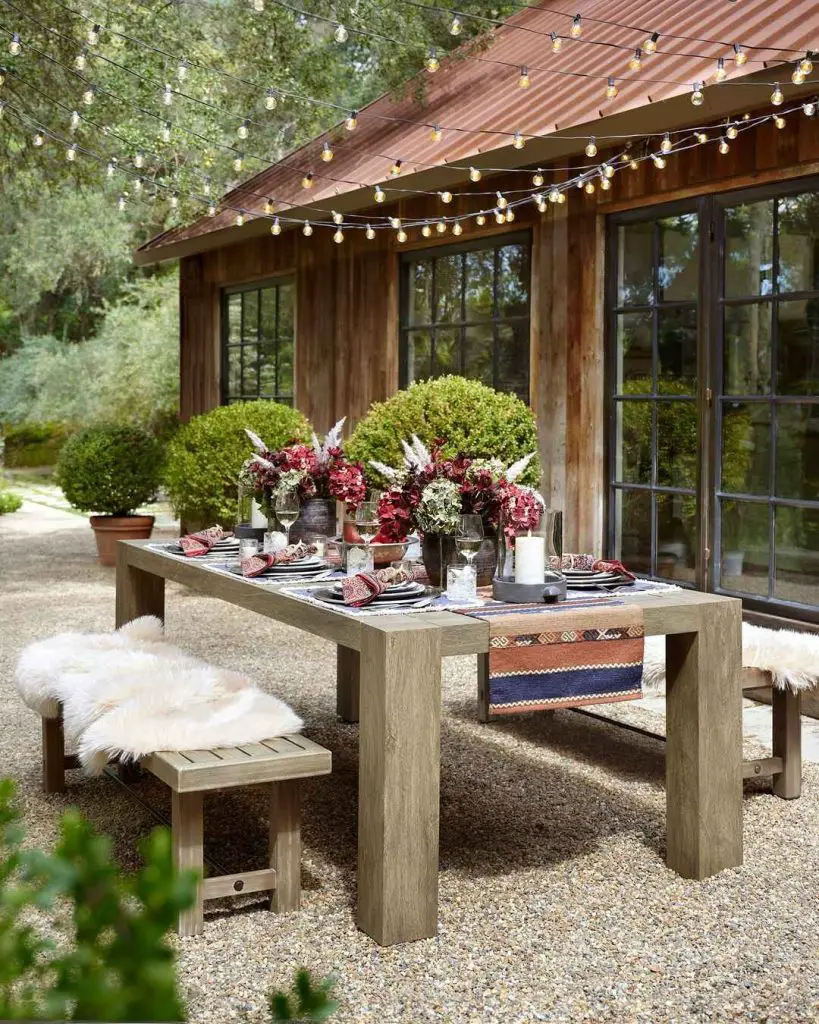 Outdoor wooden dining table with burgundy flowers sheepskin benches and string lights beneath rustic patio cover