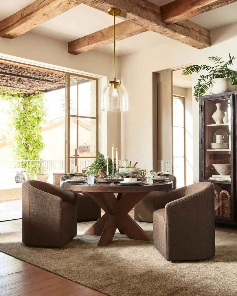 Sunlit dining room with round wooden table brown chairs and exposed ceiling beams beneath glass pendant light