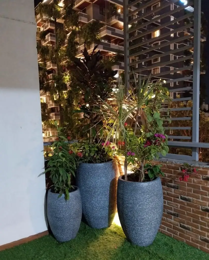Three tall grey planters with tropical plants on artificial grass balcony at night