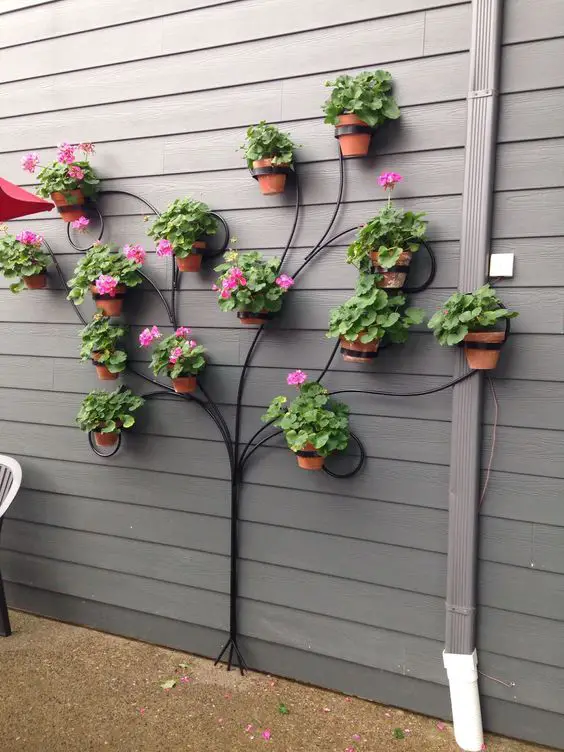Metal tree-shaped plant display on a gray wall, featuring terracotta pots with green plants and pink flowers