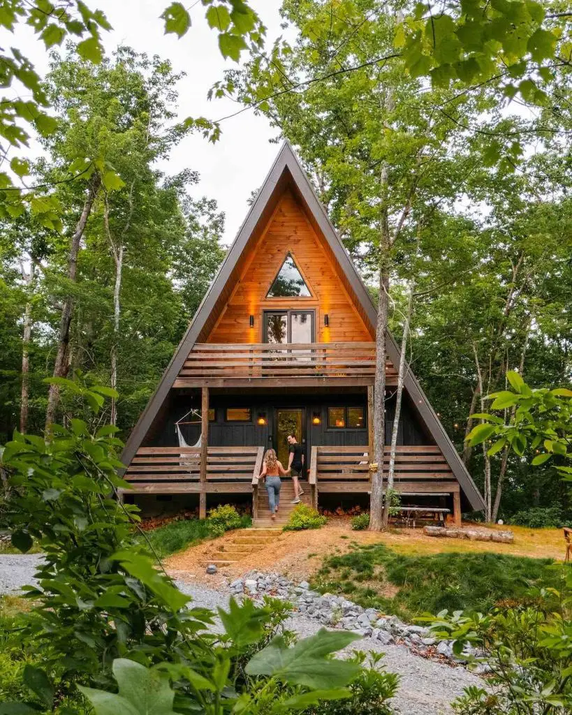 Charming A-frame cabin with wooden exterior surrounded by trees, featuring two people walking towards the entrance