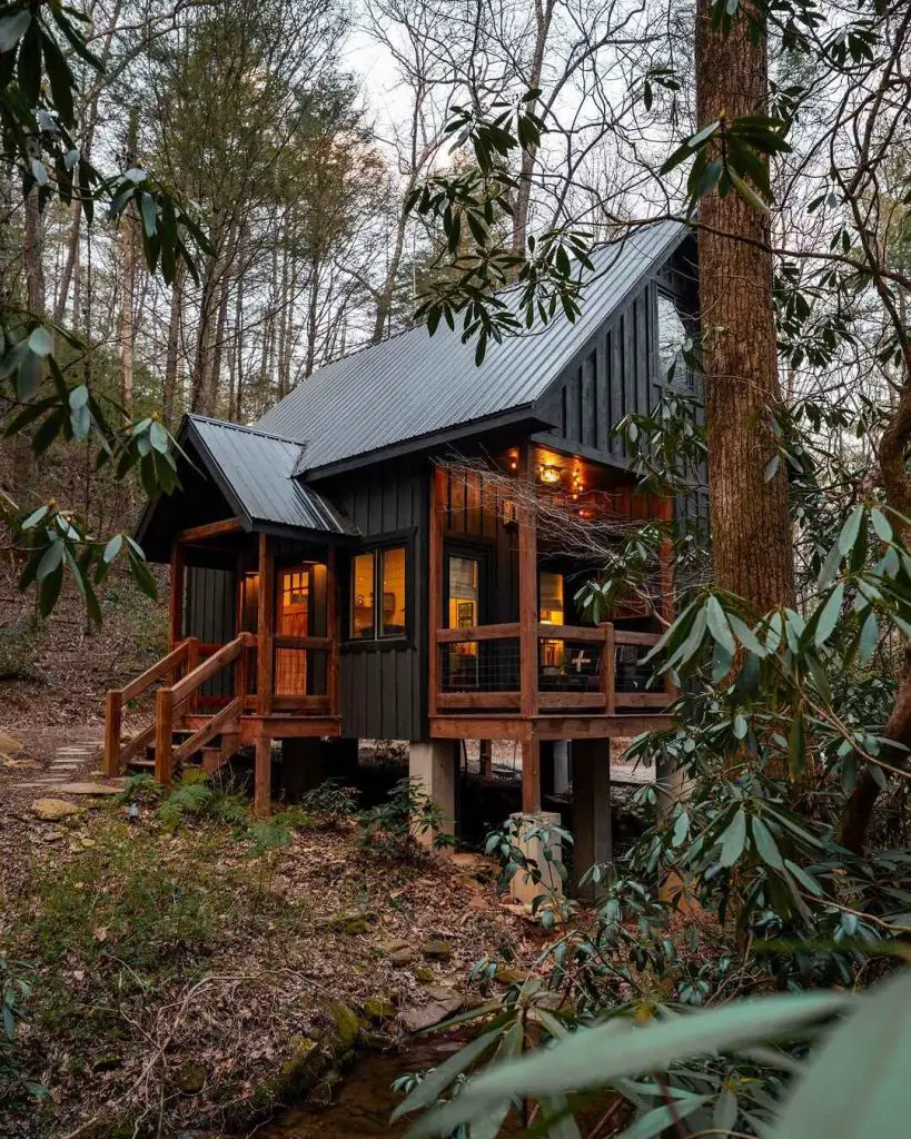 Elevated cabin with dark wooden exterior and large windows, surrounded by trees and a stream, featuring a wooden porch and steps.