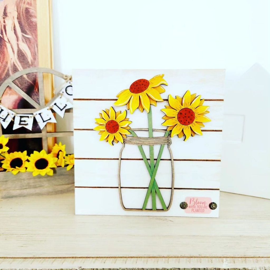Decorative piece with sunflowers in a jar and the message 'Bloom Where You Are Planted' on a wooden background