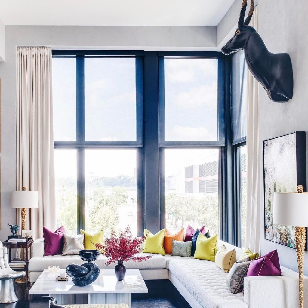 Modern living room with white L-shaped sofa, colorful pillows, and large windows overlooking a green landscape.