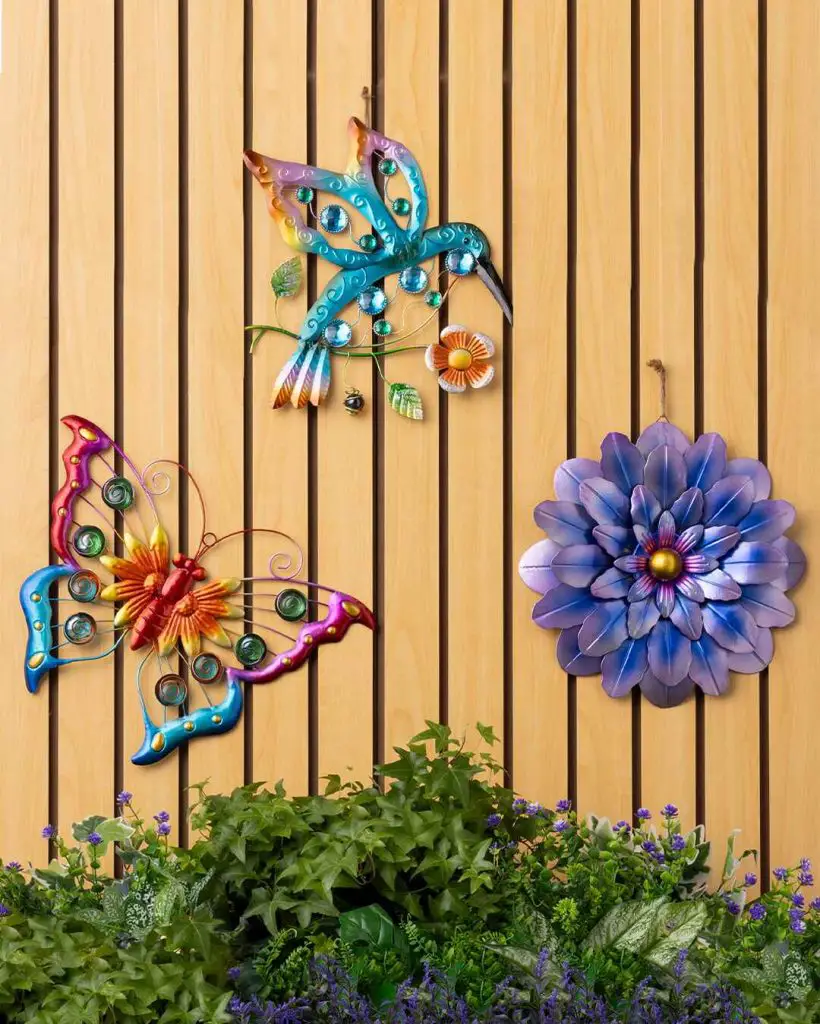Decorative metal wall art featuring a butterfly, hummingbird, and flower against a wooden backdrop with greenery below