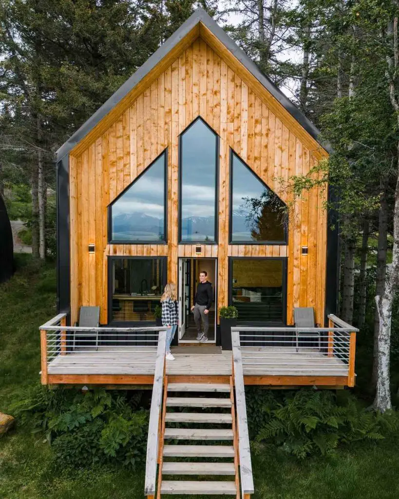 Modern A-frame cabin with large windows and a wooden deck, featuring two people at the entrance surrounded by tree