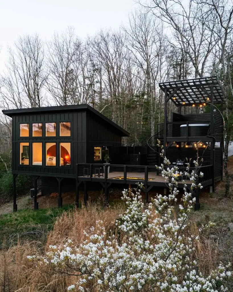 Modern black cabin with large windows and a spiral staircase, surrounded by trees and blooming flowers
