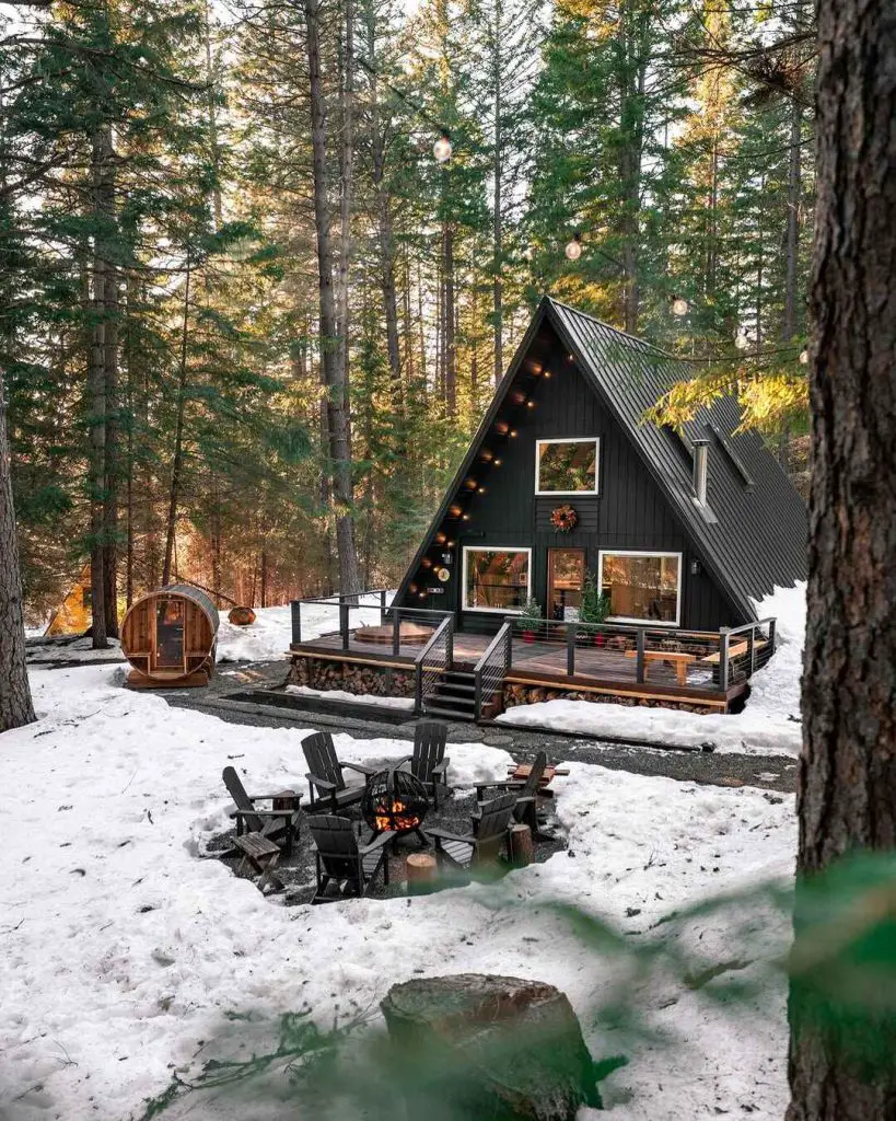 Charming black A-frame cabin surrounded by snow with a fire pit and sauna.