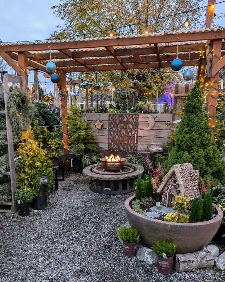 Fire pit surrounded by gravel, potted plants, and a wooden pergola with lights in a garden setting.