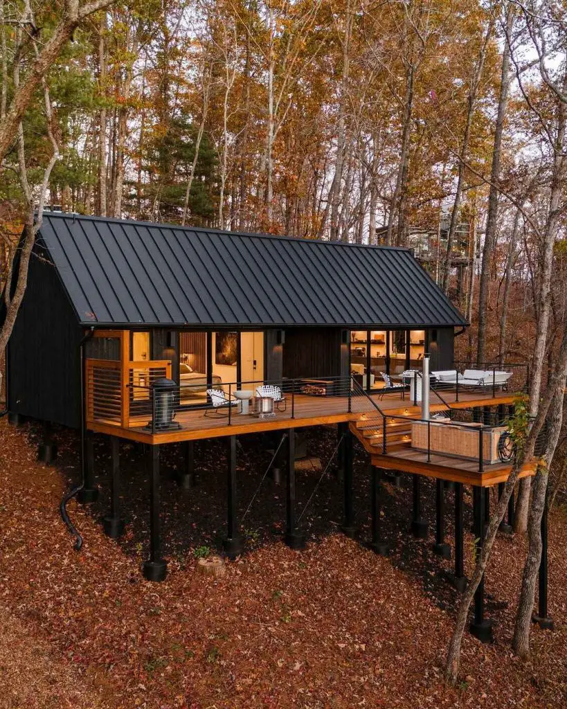 Elevated modern cabin with a black exterior and wooden deck, surrounded by autumn trees and fallen leaves