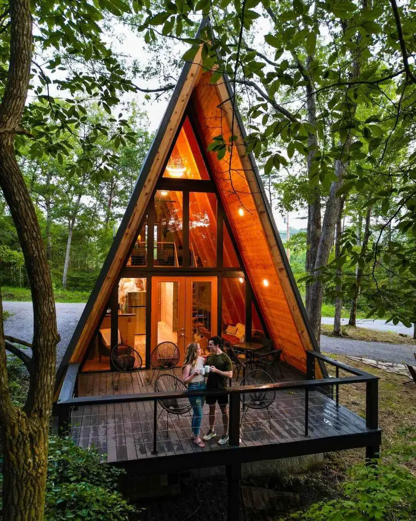 Charming A-frame cabin with large windows and a deck, surrounded by trees, featuring a couple on the porch