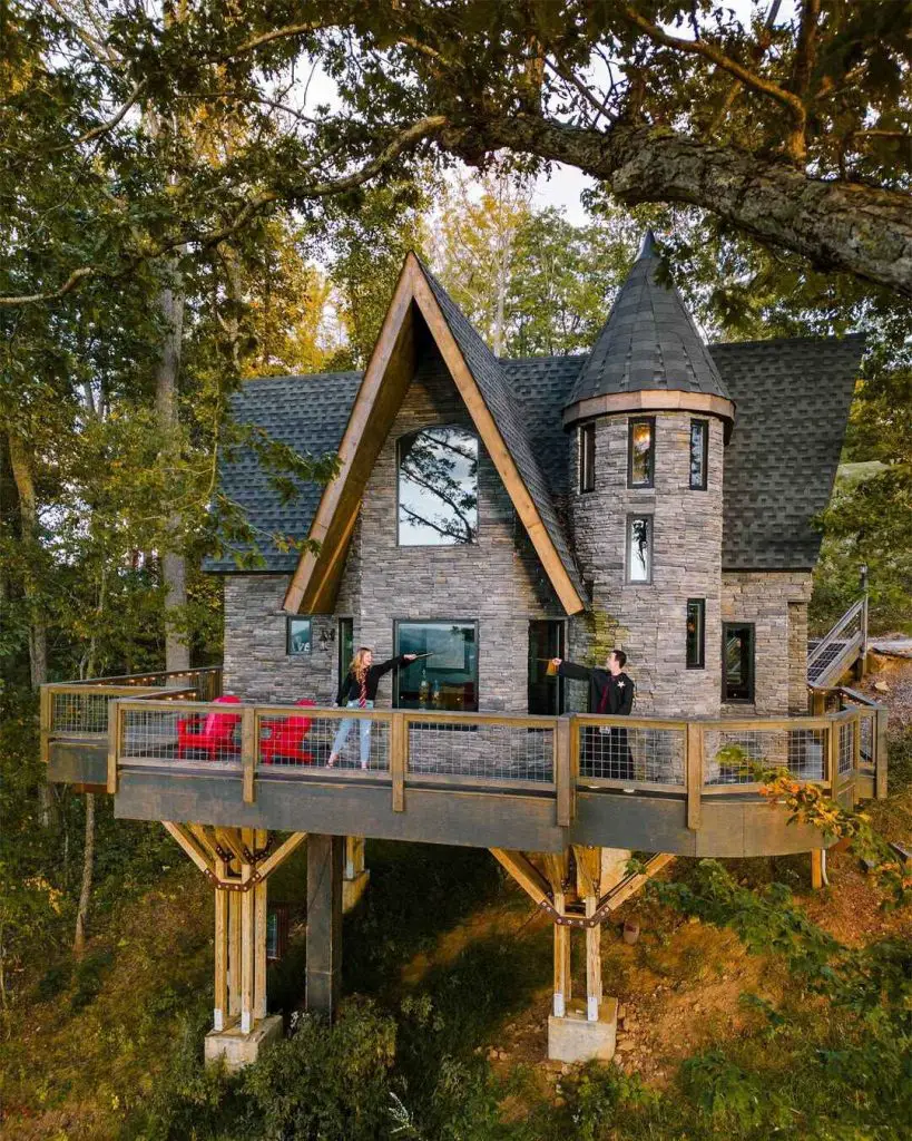 Elevated cabin with a castle-like design and large deck, surrounded by trees, featuring two people pointing on the porch