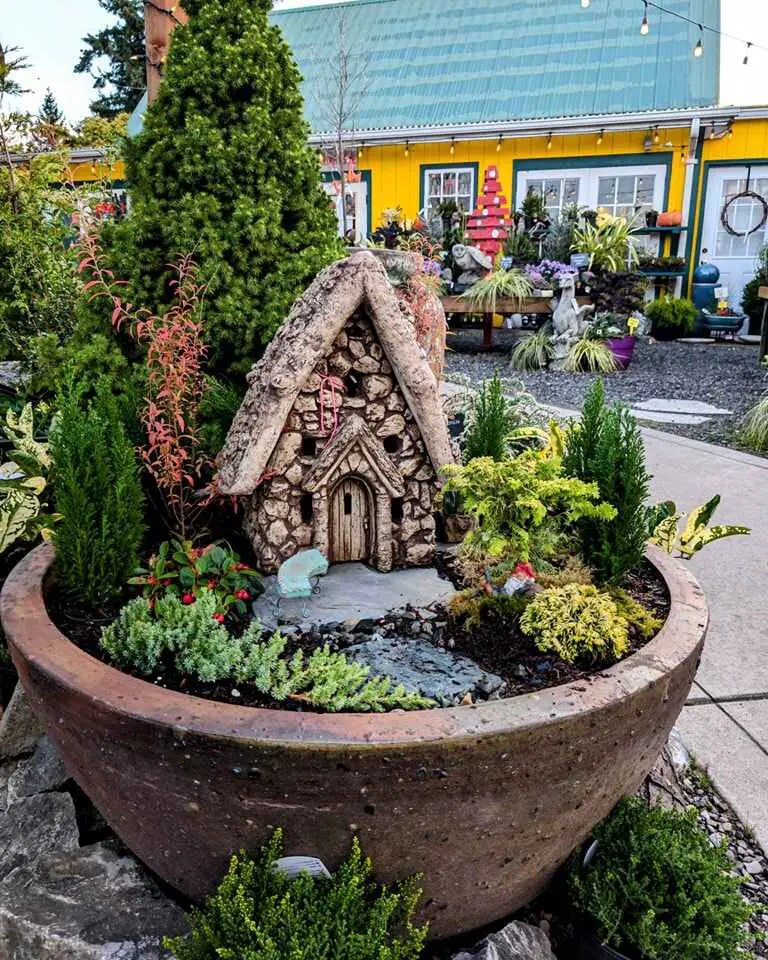 Fairy house made of natural materials in a round planter surrounded by small plants and a yellow building