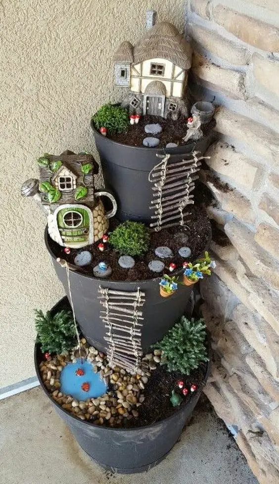 Tiered fairy garden in black pots featuring miniature houses, a pond with pebbles, and decorative plants and stones.