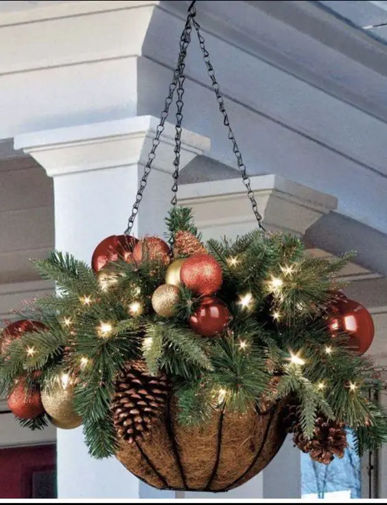 Hanging planter filled with evergreen branches, red and gold ornaments, pinecones, and twinkling lights.
